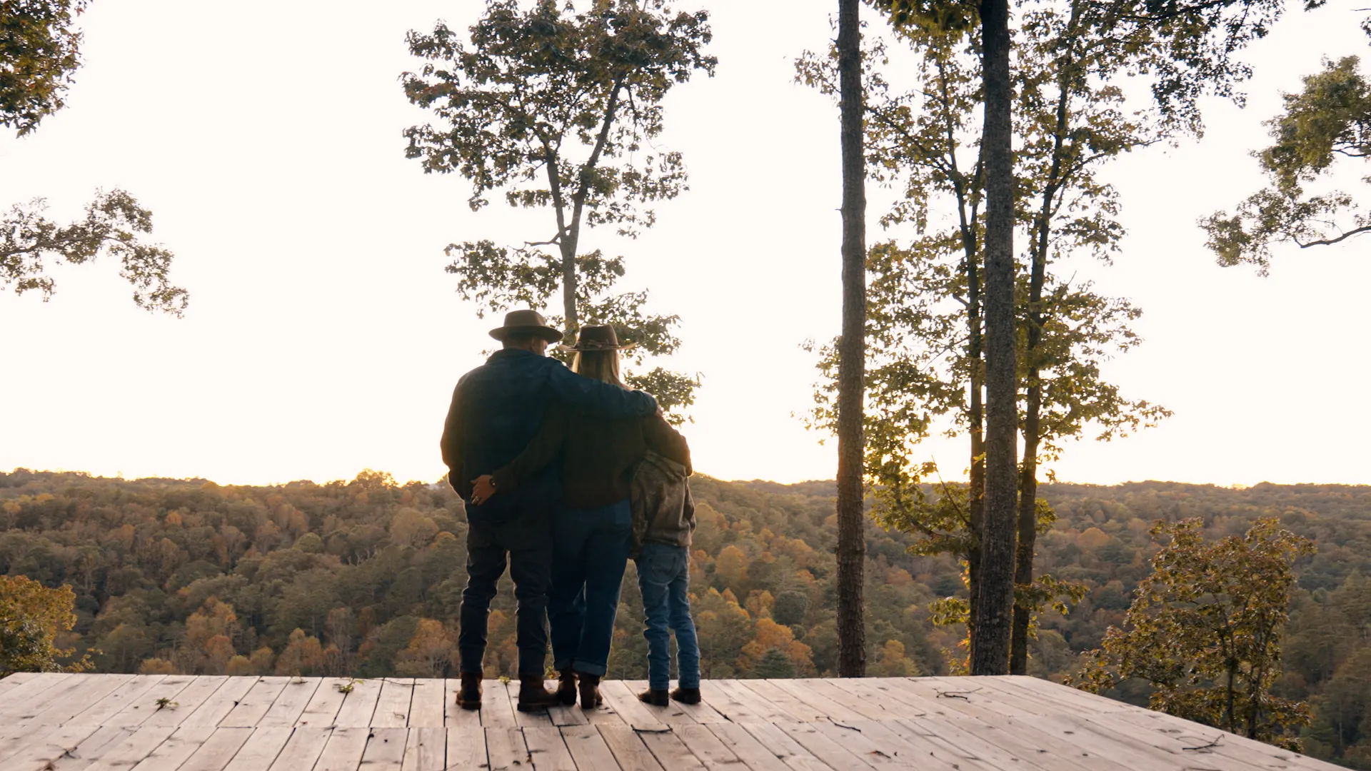 A family standing together on a wooden platform, looking out over autumn ridges at sunset